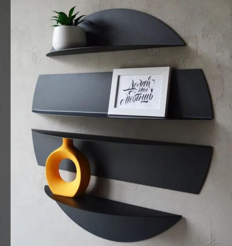 May include: A set of four dark gray metal wall shelves in a unique, semi-circular design. The shelves hold a small potted plant, a framed picture with text, and a yellow vase. The shelves are mounted on a light gray wall.