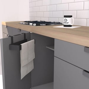 May include: A kitchen scene featuring a gray countertop with a gas stovetop and a white mug with a black rim and the word "MOMENT" printed on it. A black towel rack holds a folded gray towel. Gray cabinets and white tile complete the scene.