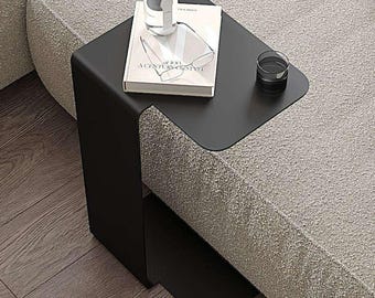 May include: A black side table with a curved design sits on a light gray couch. A book titled "Talisman of the Century" and a glass of water are on the table.