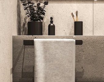 May include: A white towel hangs on a black towel bar mounted on a concrete countertop. A black planter with a green plant, a black bottle with a pump dispenser, and a black toothbrush holder with wooden toothbrushes are on the countertop.