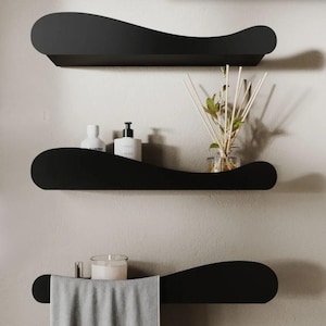 May include: Three black, wavy-edged shelves mounted on a light-colored wall. The top shelf is empty. The middle shelf holds two white bottles and a black bottle. The bottom shelf has a gray towel, a candle, and a small metal object. Home decor.