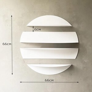 May include: White, semi-circular wall shelves arranged in a circular pattern. The shelves are 66 cm wide and 66 cm high, with a depth of 6 cm. The shelves are mounted on a gray wall. The design is modern and minimalist.
