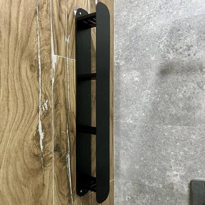 May include: A black metal shower caddy with three shelves. The caddy is mounted on a wooden tiled wall and has a sleek, modern design. The shelves are spaced apart and appear to be designed to hold toiletries.