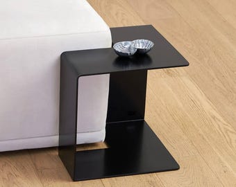 May include: A modern black side table with a unique C-shaped design. The table has a square top and base, connected by a curved support. Two decorative silver bowls sit on the tabletop. The table is positioned next to a white sofa.