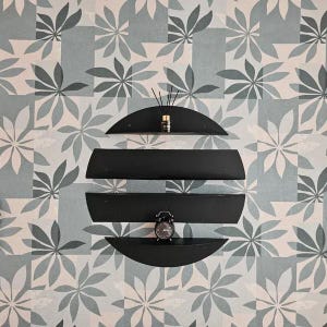 May include: A modern black wall shelf unit with three curved shelves, arranged in a circular shape. The shelves hold a small alarm clock and a fragrance diffuser. The background features a floral pattern in shades of gray and white.