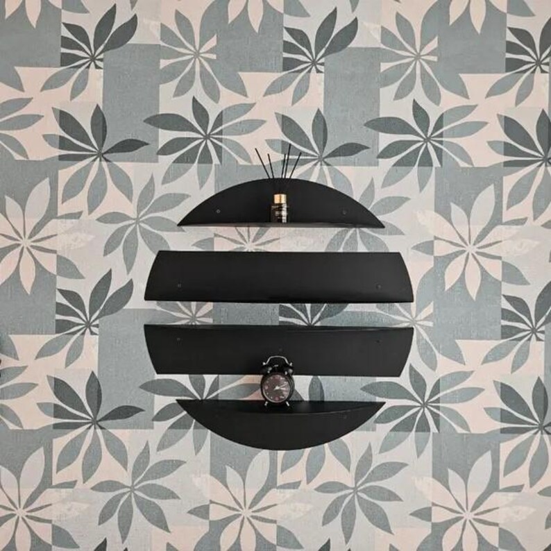 May include: A modern black wall shelf unit with three curved shelves, arranged in a circular shape. The shelves hold a small alarm clock and a fragrance diffuser. The background features a floral pattern in shades of gray and white.