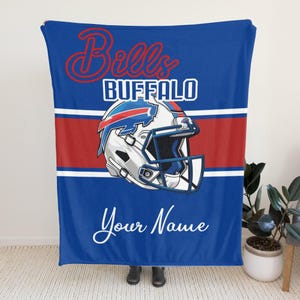 May include: Blue blanket with a red and white stripe and a Buffalo Bills football helmet graphic. The blanket has the words "Bills Buffalo" in red and white, and "Your Name" in white script.