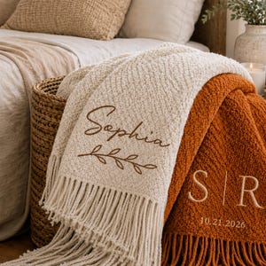 May include: A cream-coloured textured throw blanket with the name Sophia and a leaf branch embroidered in brown, alongside a rust-orange textured throw blanket featuring the initials S and R with the date 21.10.2026 embroidered in cream.