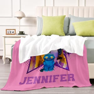 Puede incluir: Una manta de forro polar rosa con el nombre "JENNIFER" en letras moradas. La manta presenta un gato azul de dibujos animados con ojos amarillos dentro de un dise&ntilde;o en forma de estrella. La manta est&aacute; sobre una cama con almohadas blancas y verdes.