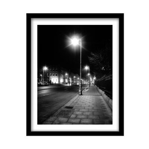 May include: A black and white photograph of a street at night with streetlights illuminating the sidewalk and buildings in the distance.
