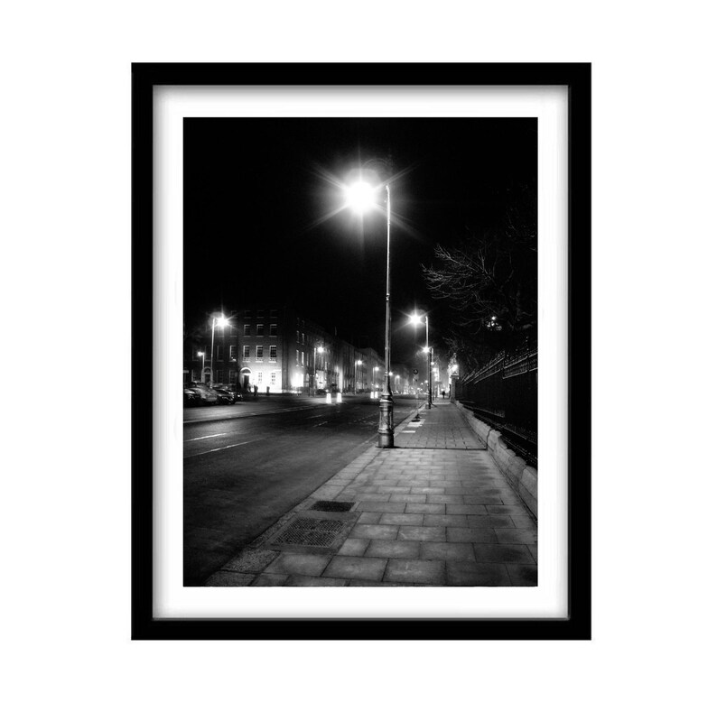 May include: A black and white photograph of a street at night with streetlights illuminating the sidewalk and buildings in the distance.