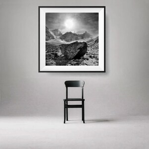 May include: Black and white framed print of a mountain landscape with a large rock in the foreground. A black chair is positioned below the framed print. The artwork depicts a dramatic sky and rugged terrain.
