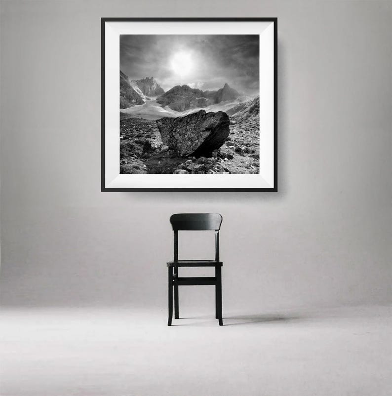 May include: Black and white framed print of a mountain landscape with a large rock in the foreground. A black chair is positioned below the framed print. The artwork depicts a dramatic sky and rugged terrain.