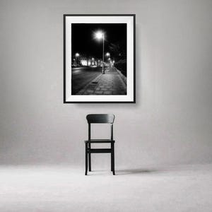 May include: Black and white photograph of a street at night with a single street lamp illuminating the sidewalk. The photo is framed and hanging on a white wall above a black chair.