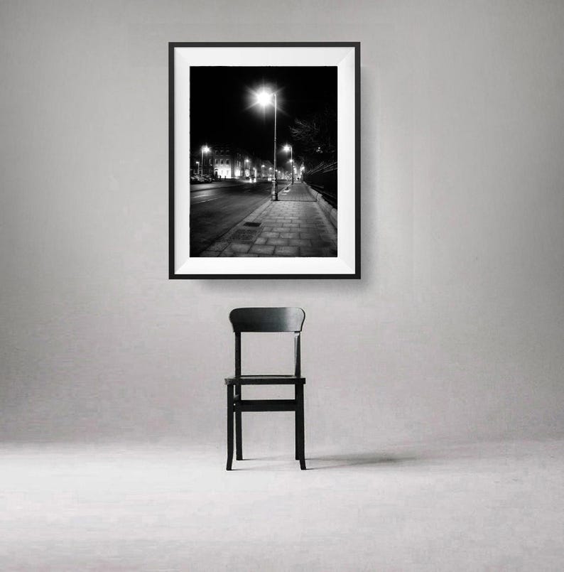 May include: Black and white photograph of a street at night with a single street lamp illuminating the sidewalk. The photo is framed and hanging on a white wall above a black chair.