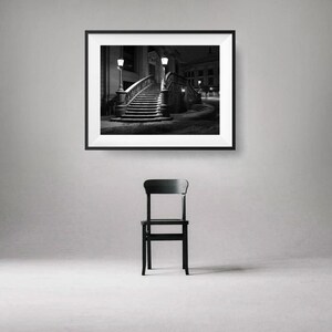 May include: A black and white framed photograph of a staircase at night, lit by street lamps. Below the photograph is a black wooden chair. The image is displayed on a gray wall.