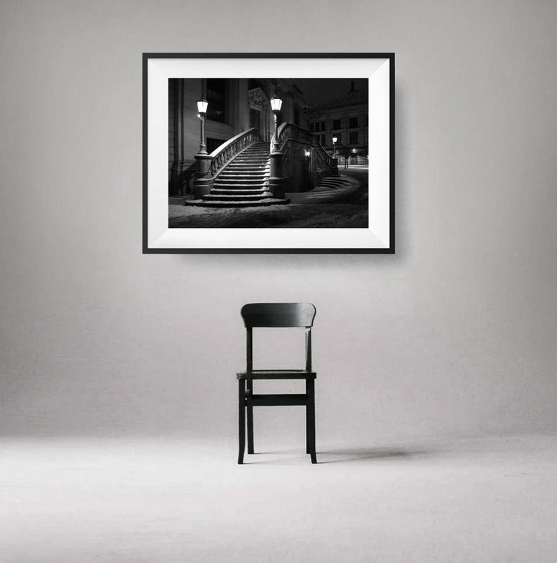 May include: A black and white framed photograph of a staircase at night, lit by street lamps. Below the photograph is a black wooden chair. The image is displayed on a gray wall.