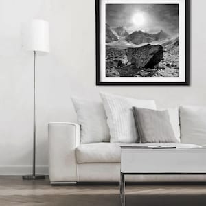 May include: Black and white framed print of a mountain landscape. The artwork features a large rock in the foreground, with mountains and a bright sky in the background. The print is displayed in a living room setting.