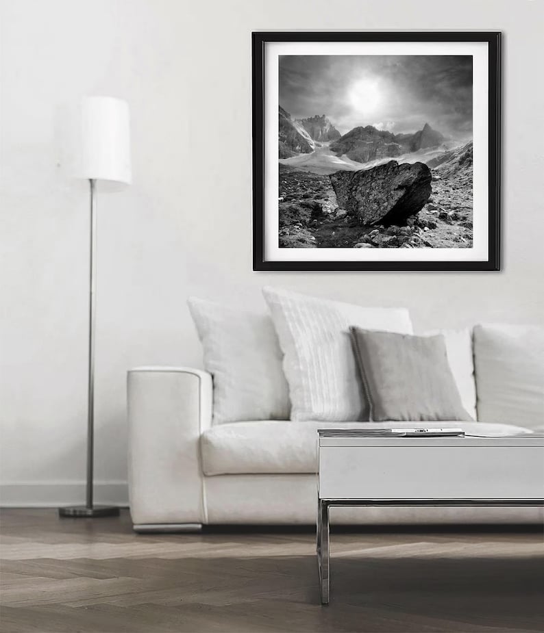 May include: Black and white framed print of a mountain landscape. The artwork features a large rock in the foreground, with mountains and a bright sky in the background. The print is displayed in a living room setting.