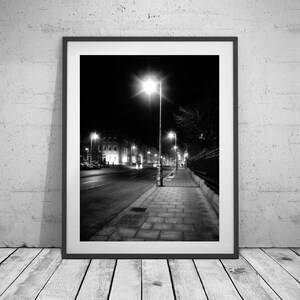 May include: A black and white photograph of a street at night with a single streetlight illuminating the sidewalk and buildings in the distance.