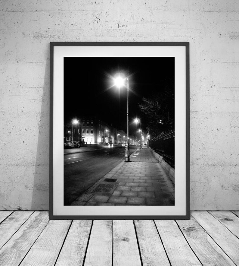 May include: A black and white photograph of a street at night with a single streetlight illuminating the sidewalk and buildings in the distance.
