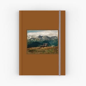 May include: A brown notebook with a landscape image of a mountain range and a wooden bench. The notebook has a brown cover and a vertical elastic band closure. The image shows a bench on a grassy hill overlooking the mountains.