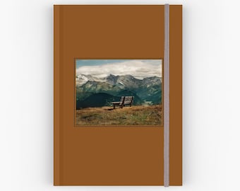May include: A brown notebook with a landscape image of a mountain range and a wooden bench. The notebook has a brown cover and a vertical elastic band closure. The image shows a bench on a grassy hill overlooking the mountains.