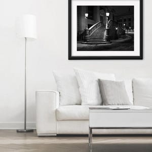 May include: Black and white framed photograph of a curved staircase with ornate railings, illuminated by street lamps. The photograph is set against a dark night sky, creating a dramatic contrast. The image is displayed above a white sofa and a modern white table.