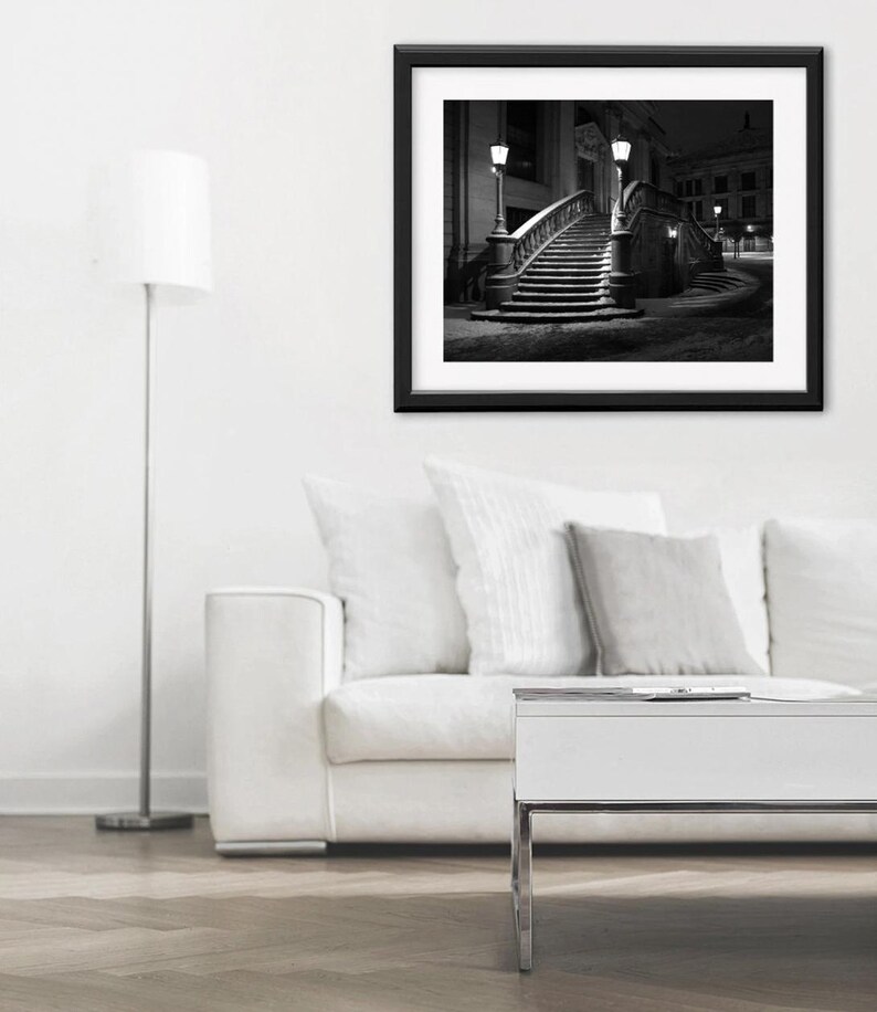 May include: Black and white framed photograph of a curved staircase with ornate railings, illuminated by street lamps. The photograph is set against a dark night sky, creating a dramatic contrast. The image is displayed above a white sofa and a modern white table.
