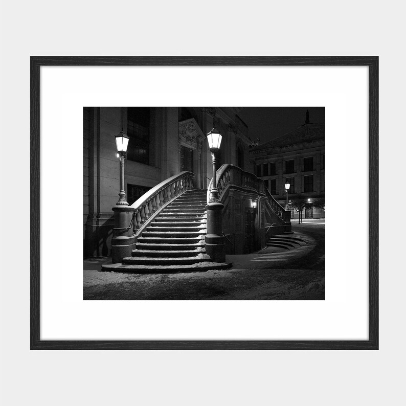 May include: Black and white framed photograph of an outdoor staircase leading up to a building. The staircase has a curved design with a stone balustrade and is flanked by ornate lampposts. Snow covers the steps and ground.
