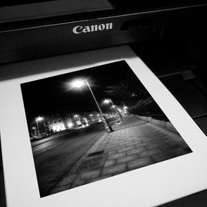 May include: A black and white photograph of a city street at night, with a streetlight illuminating the sidewalk and buildings in the distance.