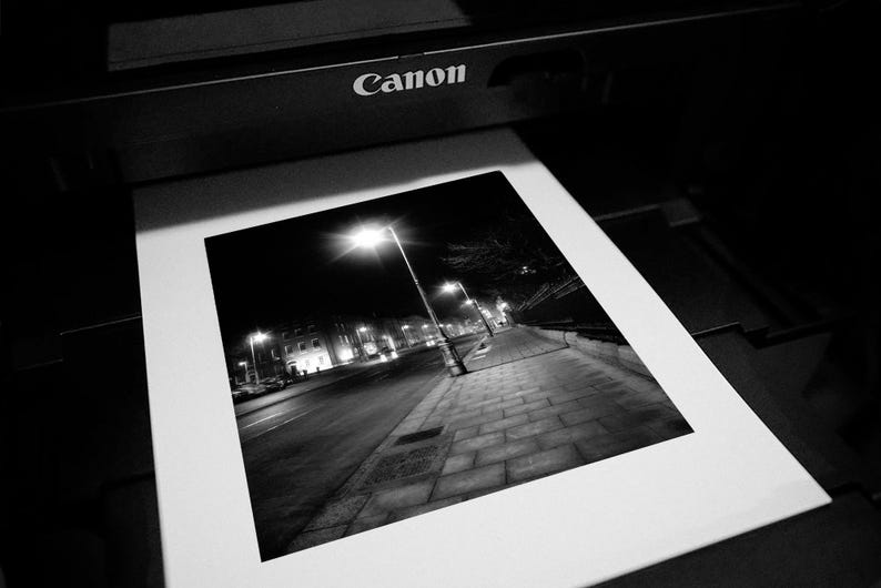 May include: A black and white photograph of a city street at night, with a streetlight illuminating the sidewalk and buildings in the distance.