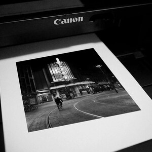Może przedstawiać: Czarno-biała fotografia sceny miejskiej drukowana przez drukarkę Canon. Obraz przedstawia osobę jadącą na rowerze ulicą z budynkiem w tle. Obraz jest wydrukowany na białym papierze.