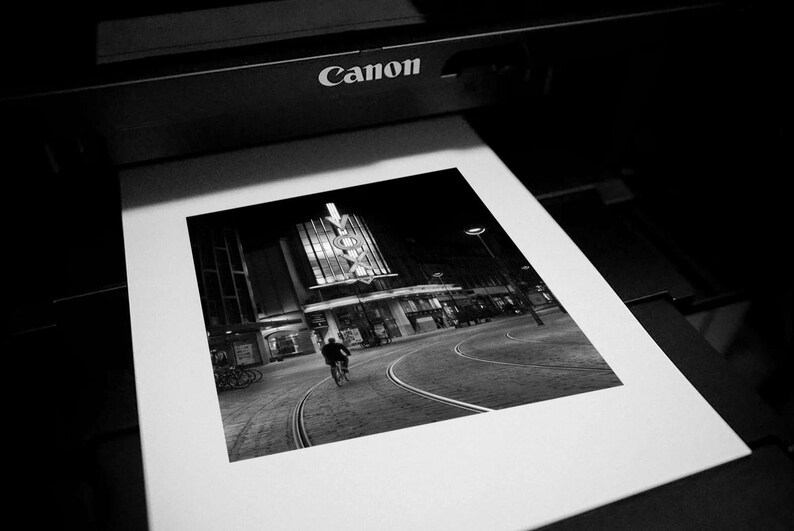 Może przedstawiać: Czarno-biała fotografia sceny miejskiej drukowana przez drukarkę Canon. Obraz przedstawia osobę jadącą na rowerze ulicą z budynkiem w tle. Obraz jest wydrukowany na białym papierze.