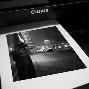May include: Black and white photograph of a printed image featuring a statue in front of a city skyline at night. The word "Canon" is visible on the printer.
