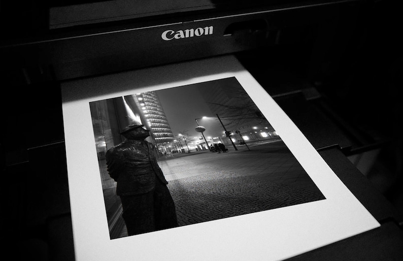 May include: Black and white photograph of a printed image featuring a statue in front of a city skyline at night. The word "Canon" is visible on the printer.