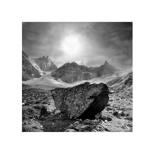 May include: Black and white landscape photograph of a mountain range under a cloudy sky. A large, textured rock sits in the foreground, with grassy terrain and smaller rocks surrounding it. The image is a study in contrasts.