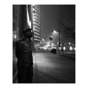 May include: Black and white photograph of a bronze statue of a man in a suit and hat, standing on a cobblestone sidewalk. The statue is looking up towards a tall, illuminated building. The background shows a city street at night with streetlights and other buildings.
