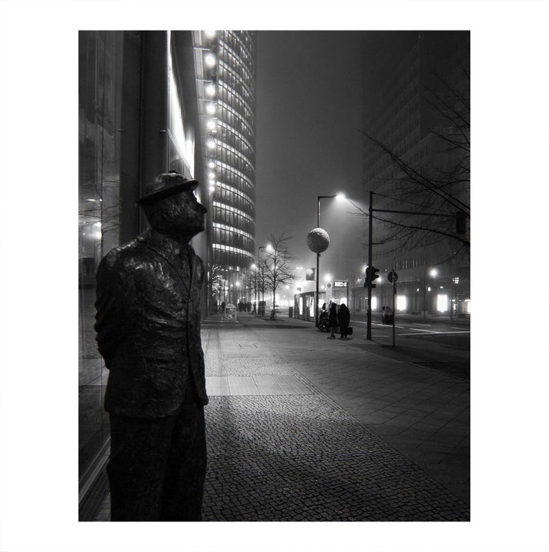 May include: Black and white photograph of a bronze statue of a man in a suit and hat, standing on a cobblestone sidewalk. The statue is looking up towards a tall, illuminated building. The background shows a city street at night with streetlights and other buildings.