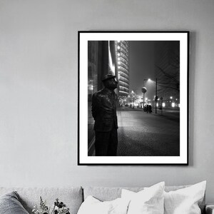 May include: Black and white framed print of a bronze statue of a man in a suit and hat, standing on a city sidewalk at night. Tall buildings and streetlights are visible in the background, creating a dramatic urban scene.