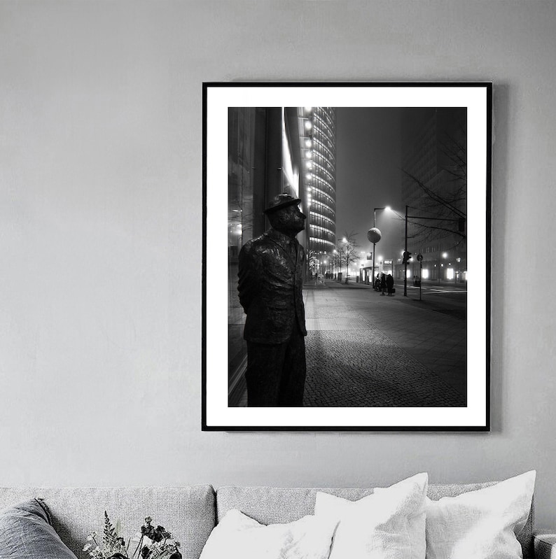 May include: Black and white framed print of a bronze statue of a man in a suit and hat, standing on a city sidewalk at night. Tall buildings and streetlights are visible in the background, creating a dramatic urban scene.