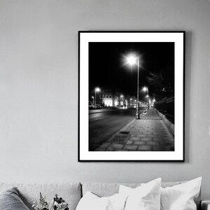 May include: Black and white photograph of a street at night with streetlights illuminating the sidewalk and buildings in the distance.