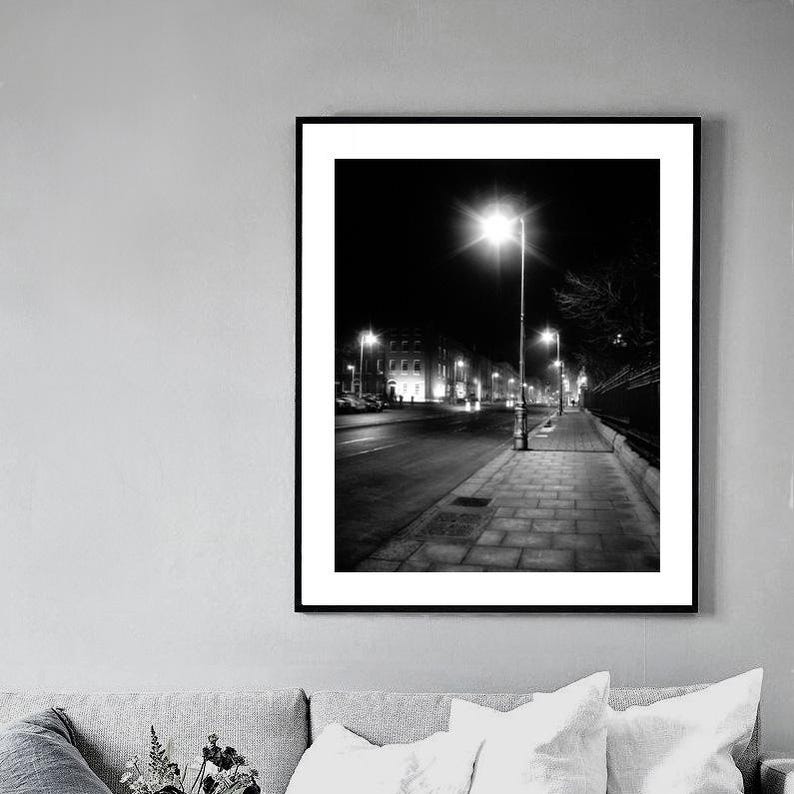 May include: Black and white photograph of a street at night with streetlights illuminating the sidewalk and buildings in the distance.