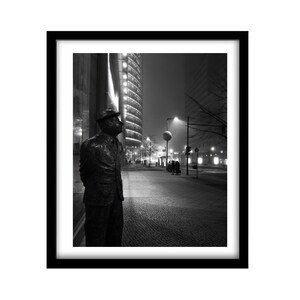 May include: Black and white photograph of a bronze statue of a man in a hat and coat, standing on a city sidewalk. The statue is in front of a modern building with a curved facade. The background shows a street with streetlights and other buildings.