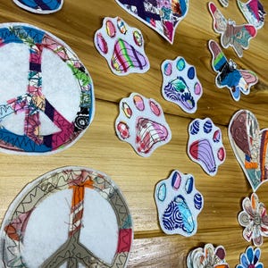 May include: A collection of fabric patches featuring various shapes including peace signs, hearts, paw prints, butterflies, and flowers. Each patch is constructed from multiple colorful fabric scraps with visible stitching and white felt backing.