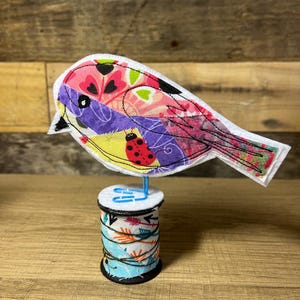 May include: A colorful patchwork bird figurine perched on a spool of thread with a blue and white patterned design.