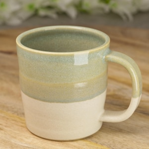 May include: A handmade ceramic mug with a two-tone design. The bottom half is a creamy white, transitioning to a light green and tan upper half. The mug has a rounded handle and is suitable for hot or cold beverages.