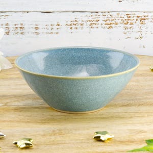May include: A speckled blue ceramic bowl with a light tan rim. The bowl is set on a wooden surface with gold and silver star-shaped decorations. A white flower is visible in the background.