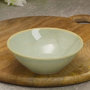 May include: A light green ceramic bowl with a speckled texture and a light yellow rim. The bowl is set on a wooden cutting board with a handle. The bowl is likely for serving food.