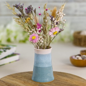 May include: A small, blue and white ceramic vase contains a bouquet of dried flowers. The vase has a light blue base, a darker blue middle section, and a white rim. The bouquet includes pink daisies, purple statice, and wheat stalks.